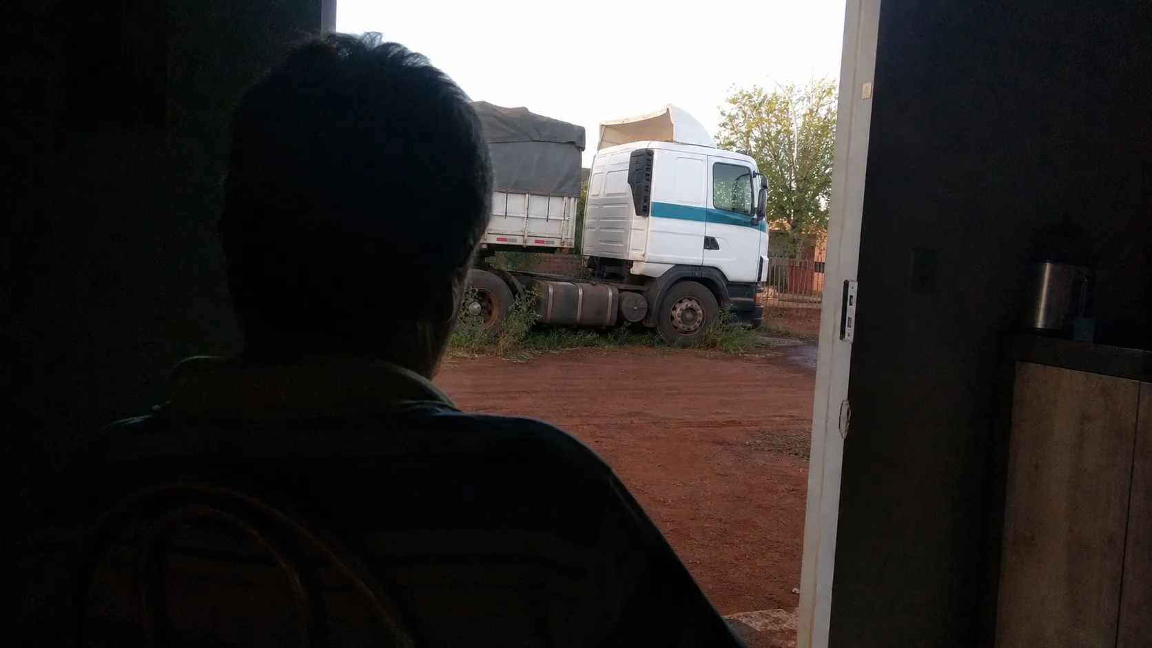 Caminhão parado no quintal de casa visto de dentro da sala, com a silhueta de um homem sentado olhando para ele
