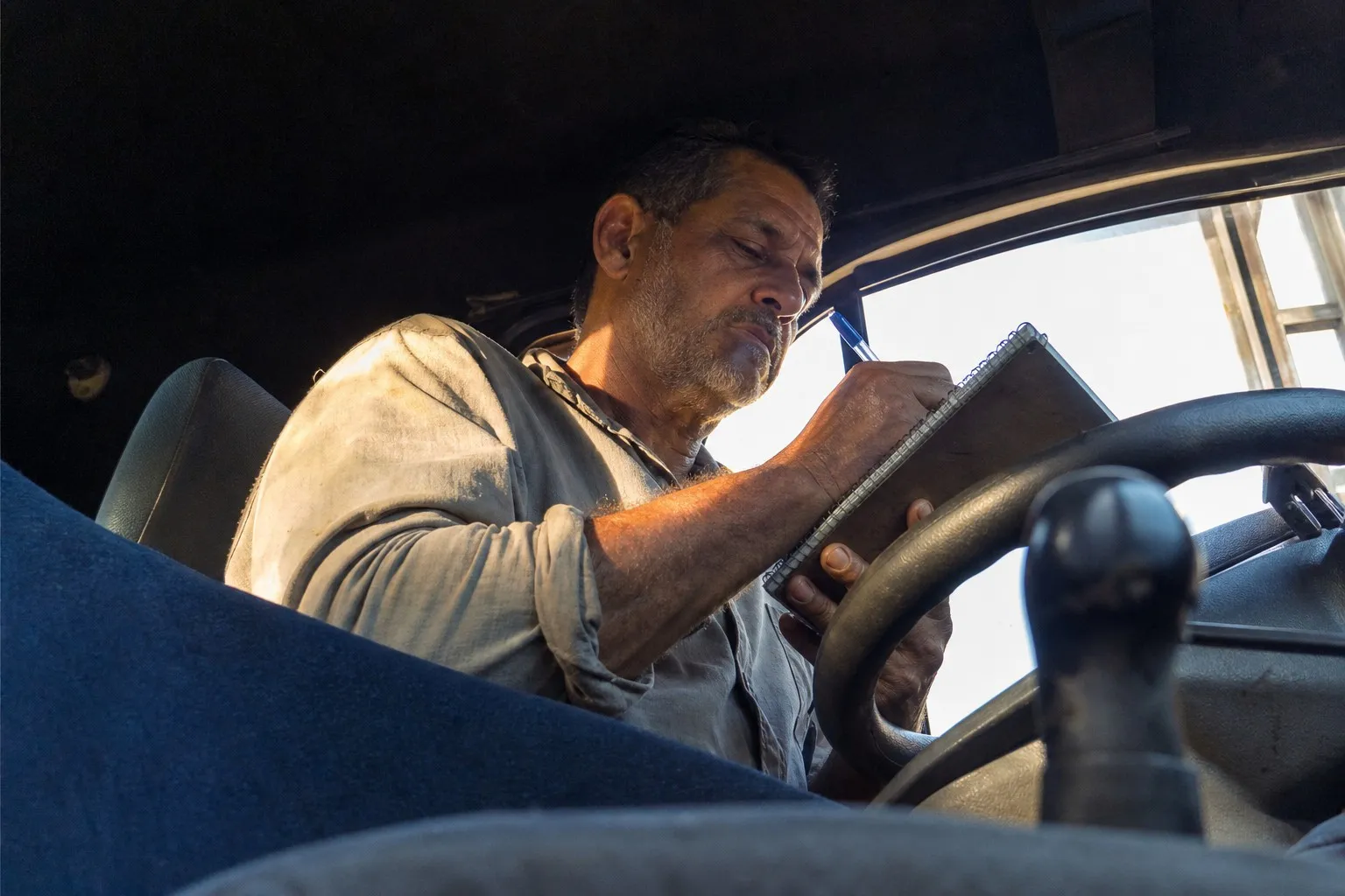 Caminhoneiro brasileiro de meia-idade sentado ao volante, visto de baixo do banco do passageiro, escrevendo com uma caneta azul num caderninho de espiral apoiado no aro do volante. A luz do fim da tarde entra pela janela do motorista e ilumina o rosto e o antebraço dele