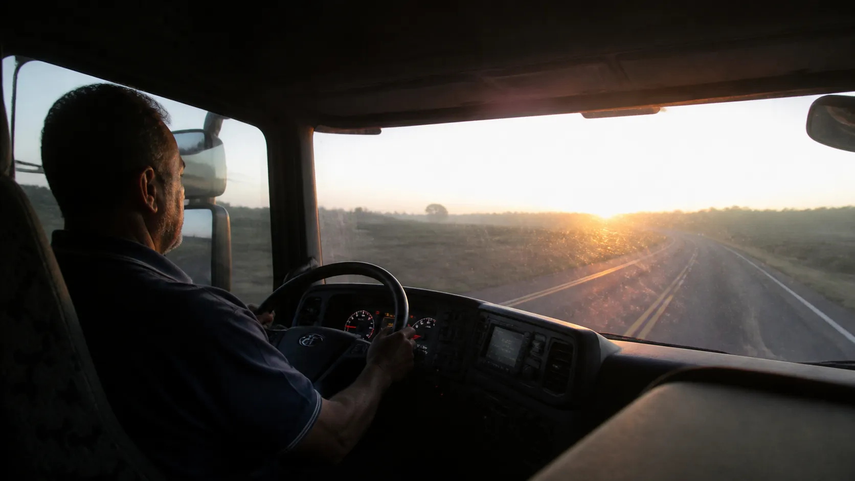 Caminhoneiro de uns cinquenta anos ao volante, dirigindo numa rodovia de pista simples ao amanhecer, com o sol baixo entrando pelo para-brisa