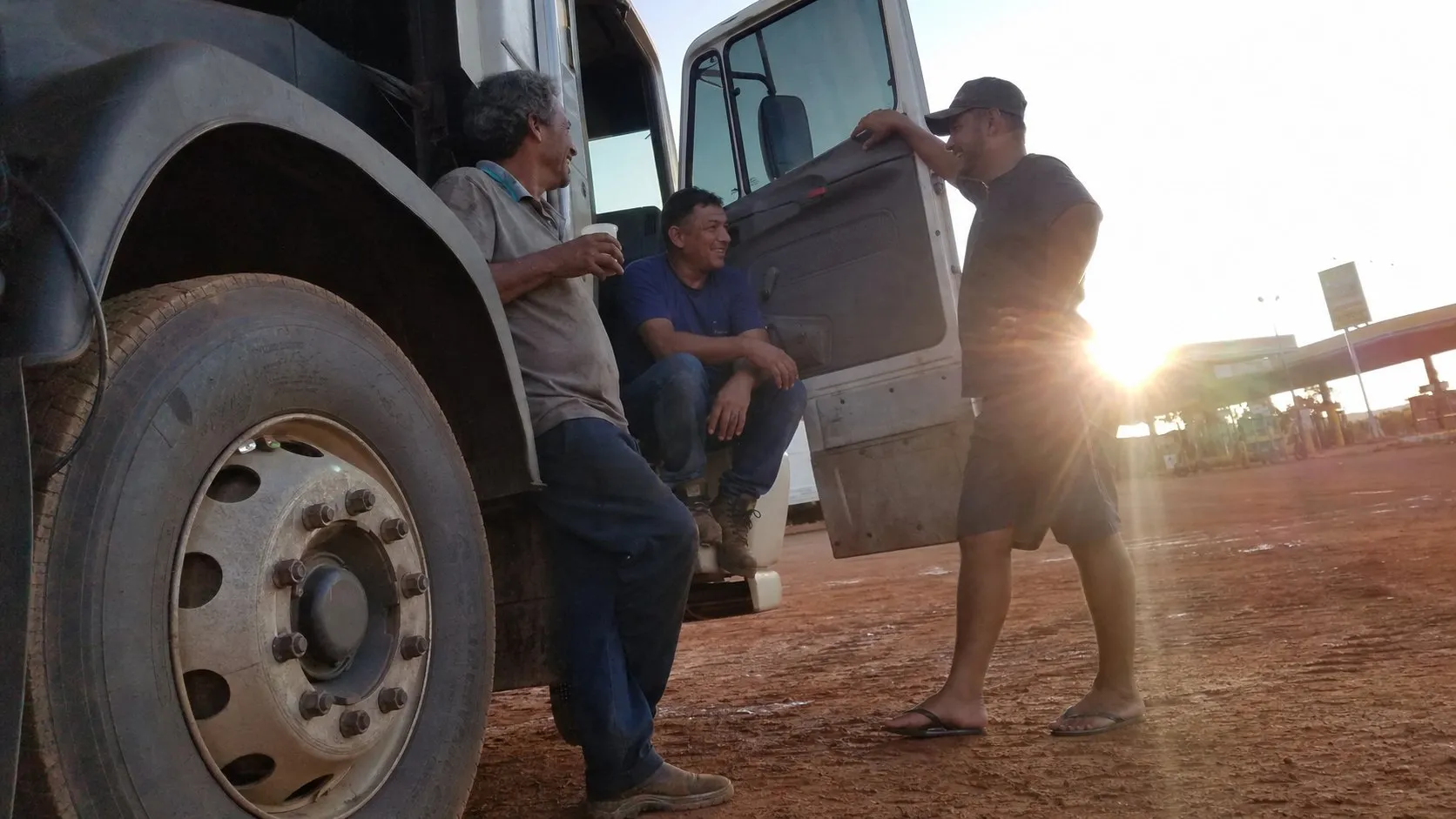 Caminhoneiro e seus dois ajudantes conversando ao lado do caminhão numa parada de estrada, de manhã cedo