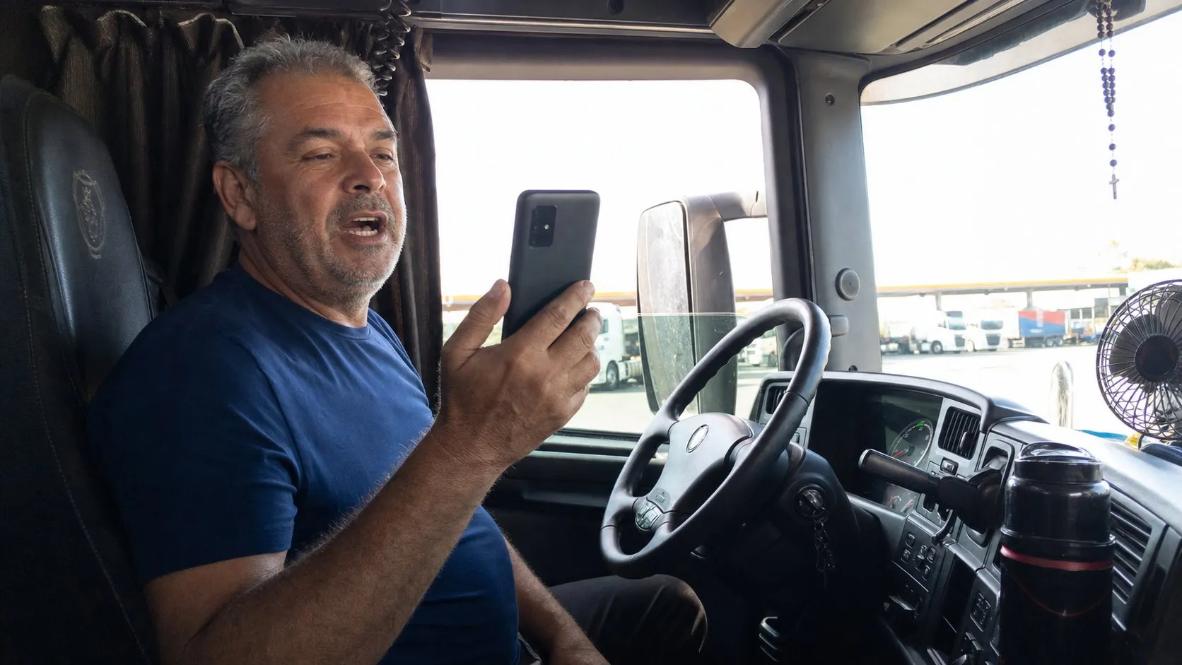 Caminhoneiro sentado na cabine do caminhão parado num posto, falando ao celular numa consulta por vídeo no fim da tarde