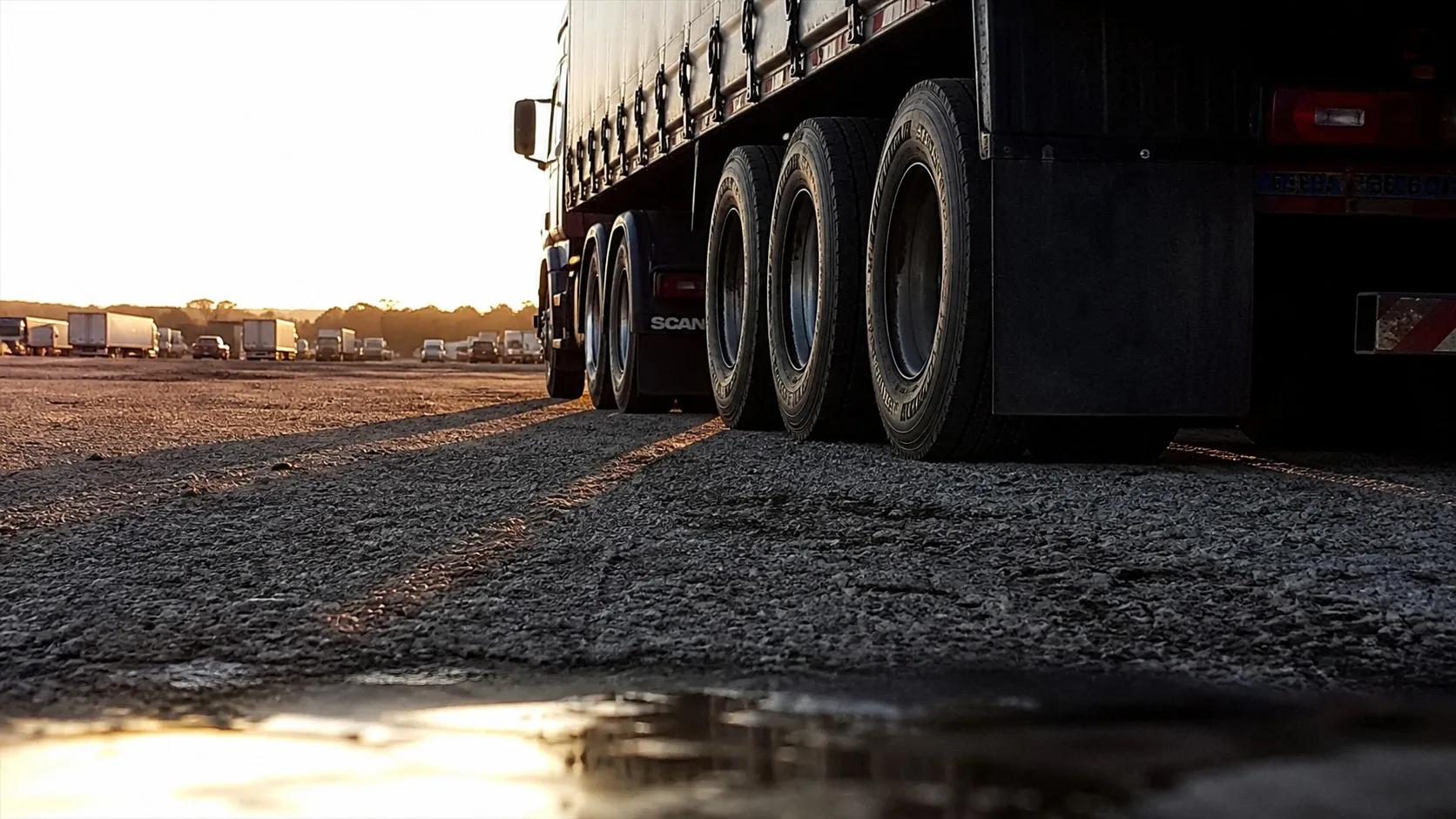 Carreta carregada e coberta com lona parada no pátio de cascalho de um posto no fim da tarde, vista de baixo, quase do chão, com as sombras longas das rodas do semirreboque se estendendo em direção à câmera e outras carretas enfileiradas ao fundo