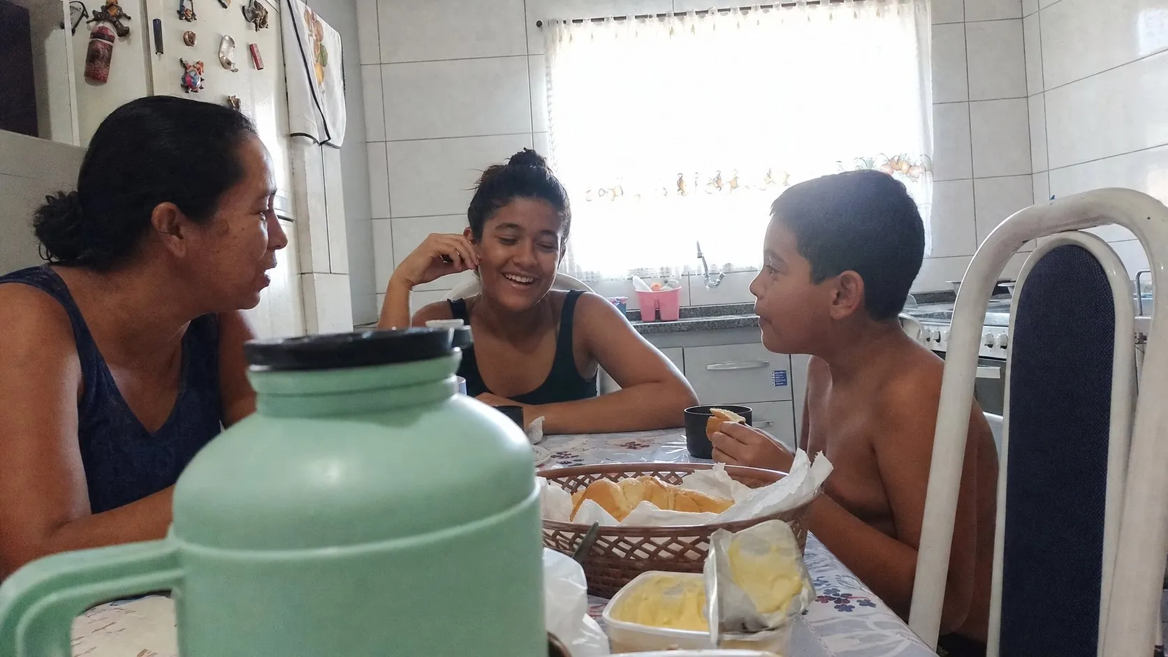 Mãe e dois filhos tomando café da manhã na cozinha de casa, com uma cadeira vazia à mesa