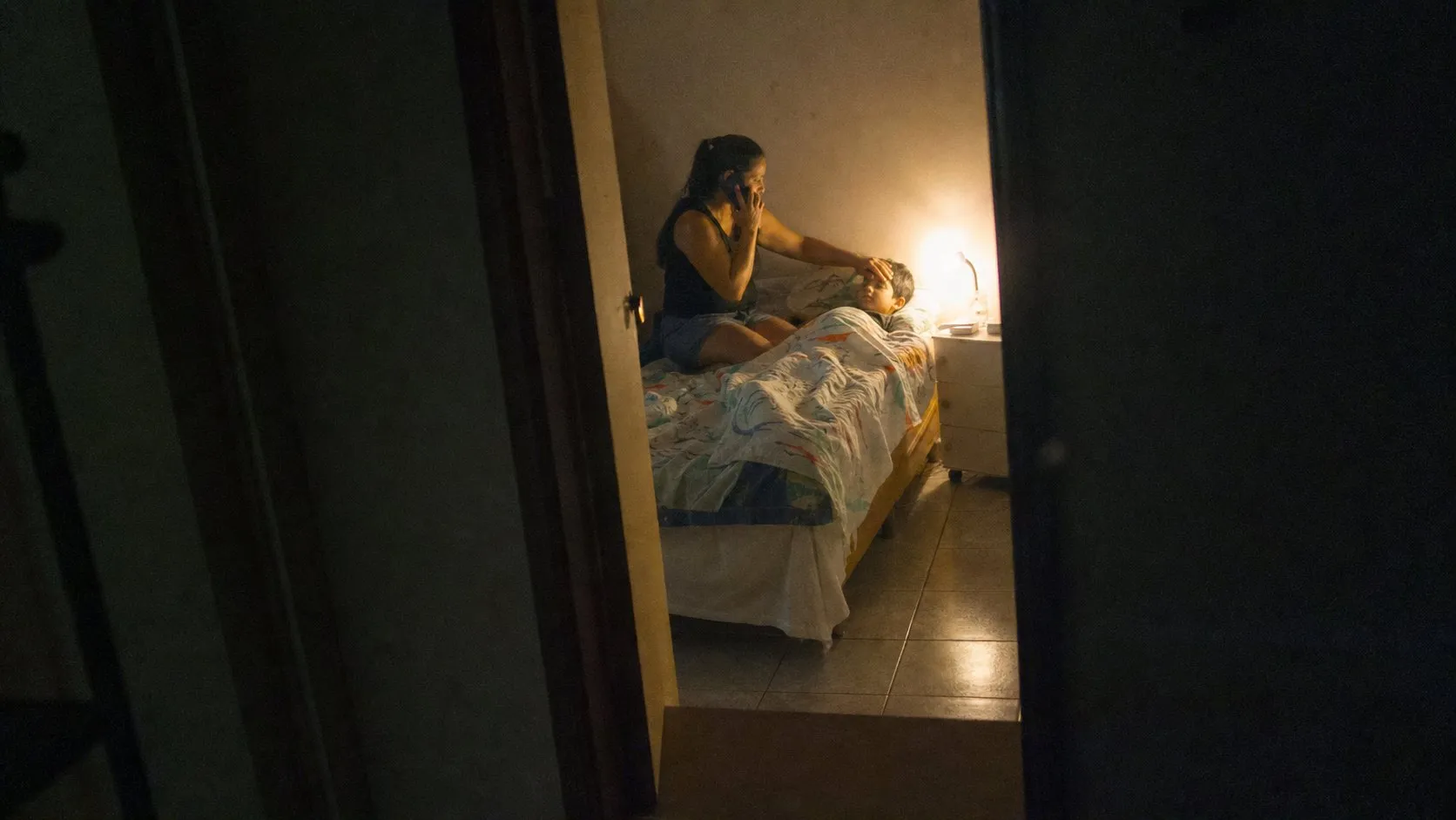 Mãe sentada na cama do filho à noite, com a mão na testa dele medindo a febre e o telefone no ouvido